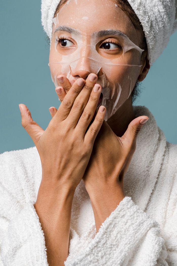 Crop surprised young African American female with towel on head and sheet mask in bathrobe covering mouth with hands and looking away during skin care routine against blue background