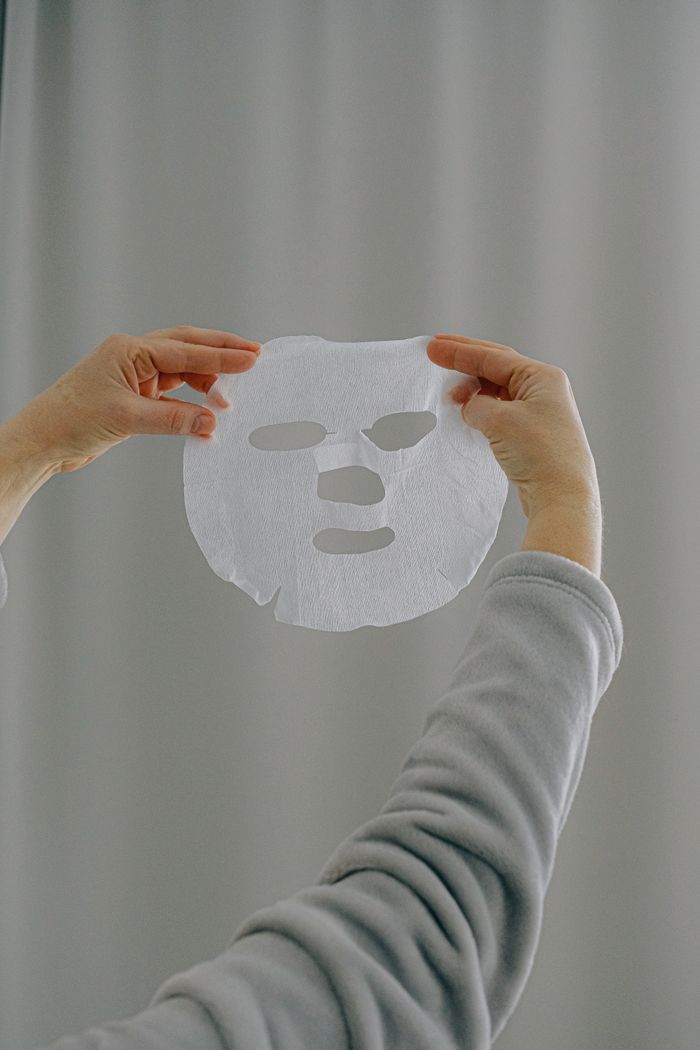 Unrecognizable female in casual clothes holding white sheet mask for face in hands against gray background in daytime at home