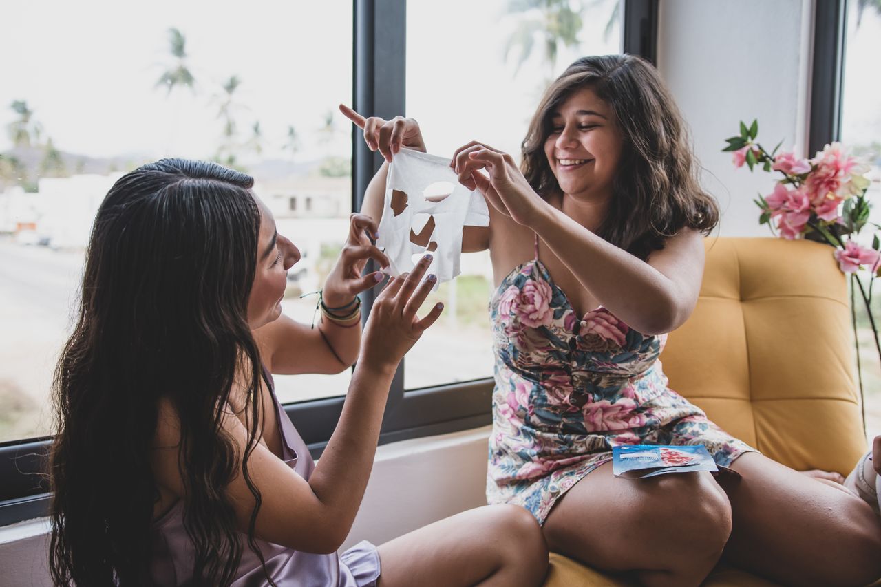 Two young women applying sheet masks while bonding indoors on a sunny day.